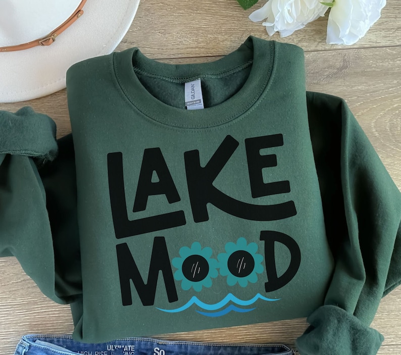 May include: A dark green sweatshirt with the text "LAKE MOOD" in black lettering. The text is stylized with a wavy line underneath and two cartoon eyes with eyelashes.