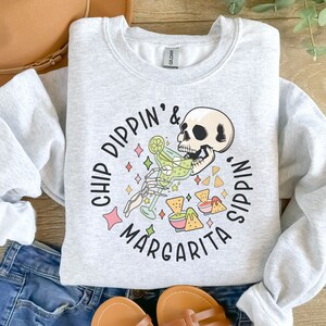 May include: Gray sweatshirt with a graphic of a skeleton drinking a margarita and eating chips. The text reads "Chip Dippin' & Margarita Sippin'."