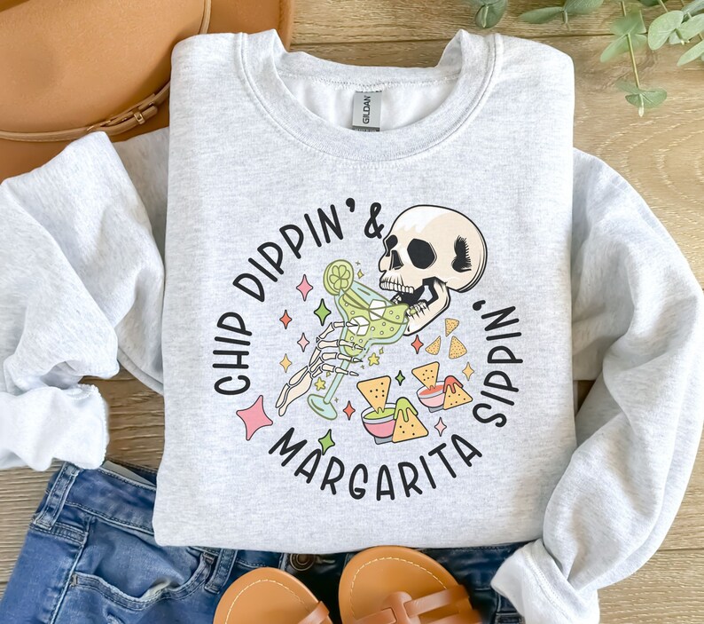 May include: Gray sweatshirt with a graphic of a skeleton drinking a margarita and eating chips. The text reads "Chip Dippin' & Margarita Sippin'."