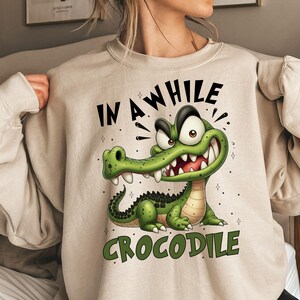 May include: Beige sweatshirt featuring a cartoon crocodile with the text "IN A WHILE CROCODILE" in black and green. The crocodile is green with white teeth and a black spine. The sweatshirt has long sleeves.