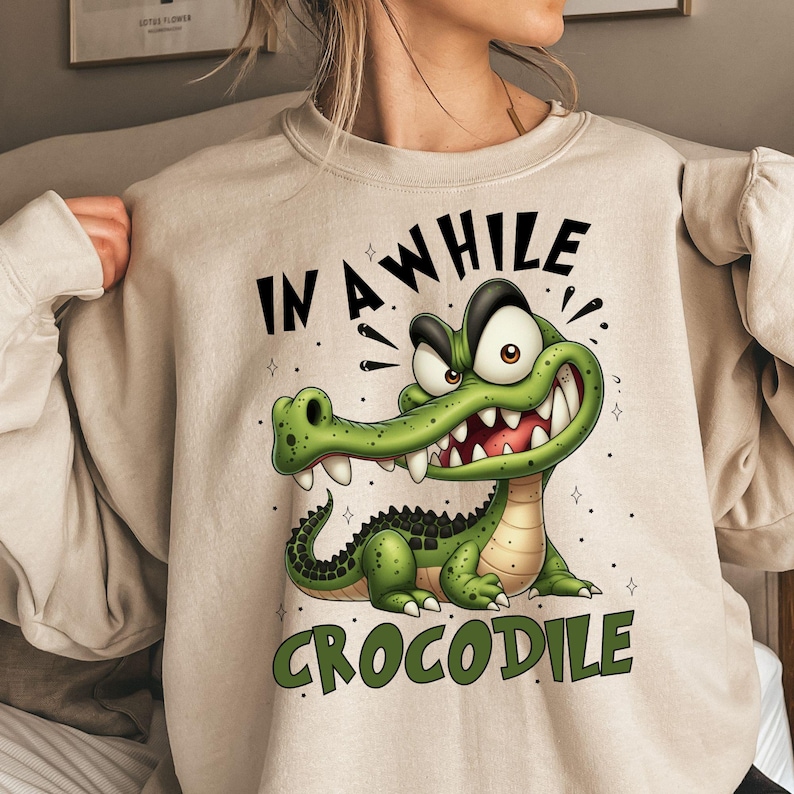 May include: Beige sweatshirt featuring a cartoon crocodile with the text "IN A WHILE CROCODILE" in black and green. The crocodile is green with white teeth and a black spine. The sweatshirt has long sleeves.