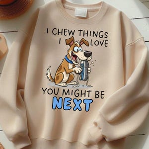 May include: A beige sweatshirt with a cartoon dog graphic chewing a blue shoe. The text reads "I CHEW THINGS I LOVE YOU MIGHT BE NEXT" in black and blue. The sweatshirt has long sleeves and a crew neck.