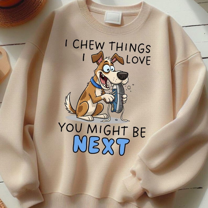 May include: A beige sweatshirt with a cartoon dog graphic chewing a blue shoe. The text reads "I CHEW THINGS I LOVE YOU MIGHT BE NEXT" in black and blue. The sweatshirt has long sleeves and a crew neck.