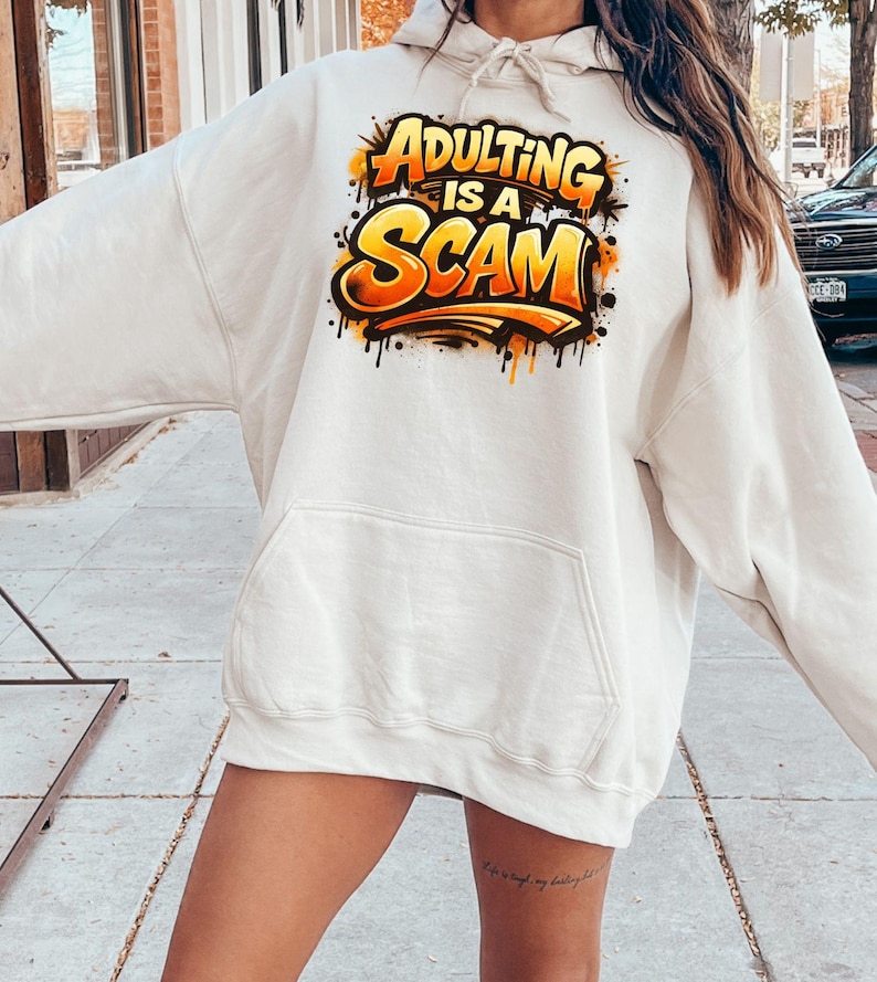 Pu&ograve; includere: Felpa con cappuccio color crema con tasca frontale e cappuccio con coulisse. La felpa presenta la scritta "Adulting is a Scam" in un design in stile graffiti con lettere arancioni e gialle, delineate in nero.