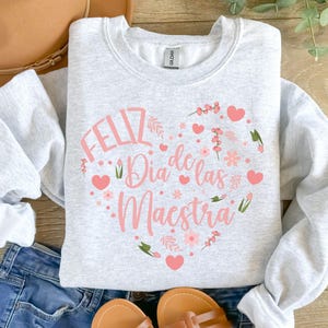 Pode incluir: Camisola cinzenta com um desenho de cora&ccedil;&atilde;o rosa que diz "Feliz Dia de las Maestra" em letras cor-de-rosa.