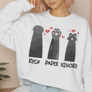 May include: White sweatshirt with a graphic of three cat paws playing rock, paper, scissors. The paws are black with pink accents. The text "Rock Paper Scissors" is printed below the paws.