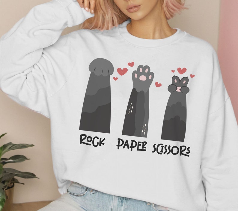 May include: White sweatshirt with a graphic of three cat paws playing rock, paper, scissors. The paws are black with pink accents. The text "Rock Paper Scissors" is printed below the paws.