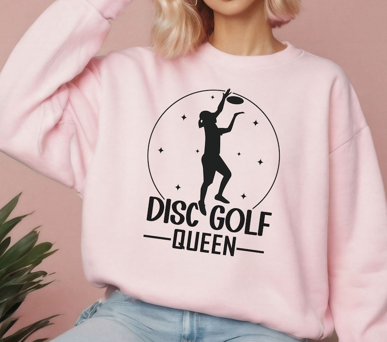 May include: Pink sweatshirt with a black graphic of a person throwing a disc, a circle with stars, and the text "Disc Golf -Queen-"