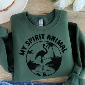 Puede incluir: Una sudadera verde oscuro con el texto "My Spirit Animal" en letras negras y una silueta negra de un flamenco de pie en el agua con palmeras en el fondo.