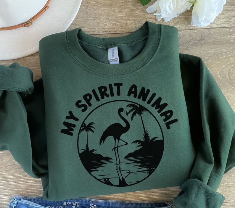 Puede incluir: Una sudadera verde oscuro con el texto "My Spirit Animal" en letras negras y una silueta negra de un flamenco de pie en el agua con palmeras en el fondo.