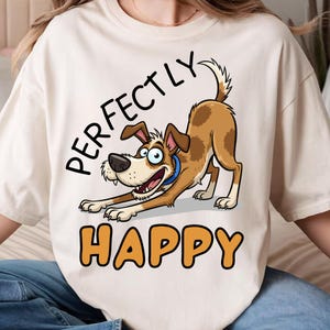 May include: Cream-colored t-shirt featuring a cartoon dog in a playful pose. The dog is brown and white with a blue collar. The words "PERFECTLY HAPPY" are printed above and below the dog in black and orange lettering.