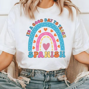 Pode incluir: Camiseta branca com um gr&aacute;fico de arco-&iacute;ris e o texto "&Eacute; um bom dia para ensinar espanhol".