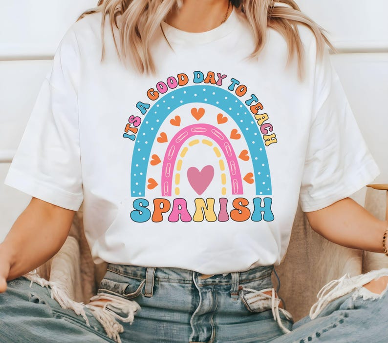Pode incluir: Camiseta branca com um gr&aacute;fico de arco-&iacute;ris e o texto "&Eacute; um bom dia para ensinar espanhol".