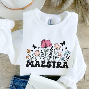 Pode incluir: Camisola branca com um design floral e a palavra "MAESTRA" em letras pretas.