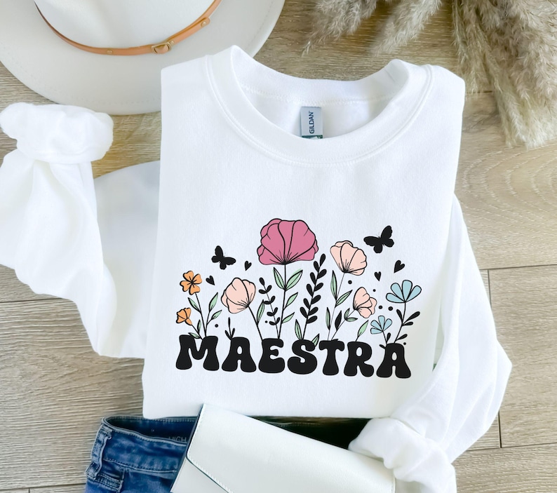 Pode incluir: Camisola branca com um design floral e a palavra "MAESTRA" em letras pretas.