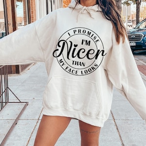 K&ouml;nnte beinhalten: Ein wei&szlig;er Hoodie mit einem schwarzen Kreis-Motiv, auf dem "I Promise I'm Nicer Than My Face Looks" steht.