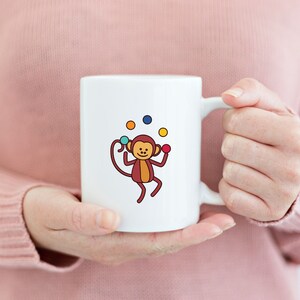 May include: White ceramic mug with a cartoon monkey juggling colorful balls. The monkey is brown with a yellow belly and has a red nose. The balls are red, yellow, blue, and green.