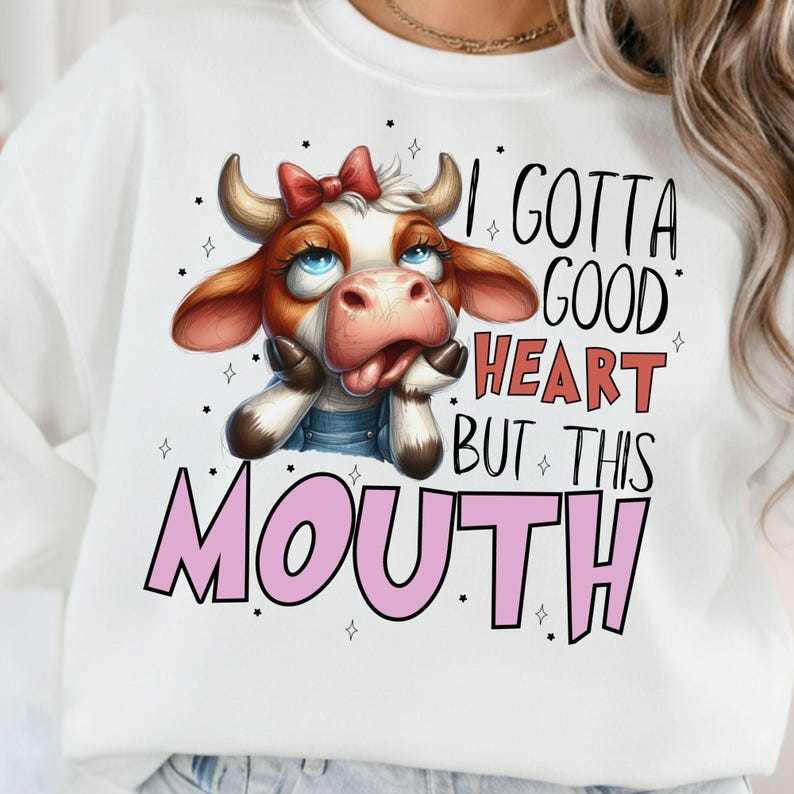 May include: White sweatshirt featuring a cartoon cow with a red bow, blue eyes, and a pink tongue. The text "I Gotta Good Heart But This Mouth" is printed in black and pink. The cow is wearing denim overalls.