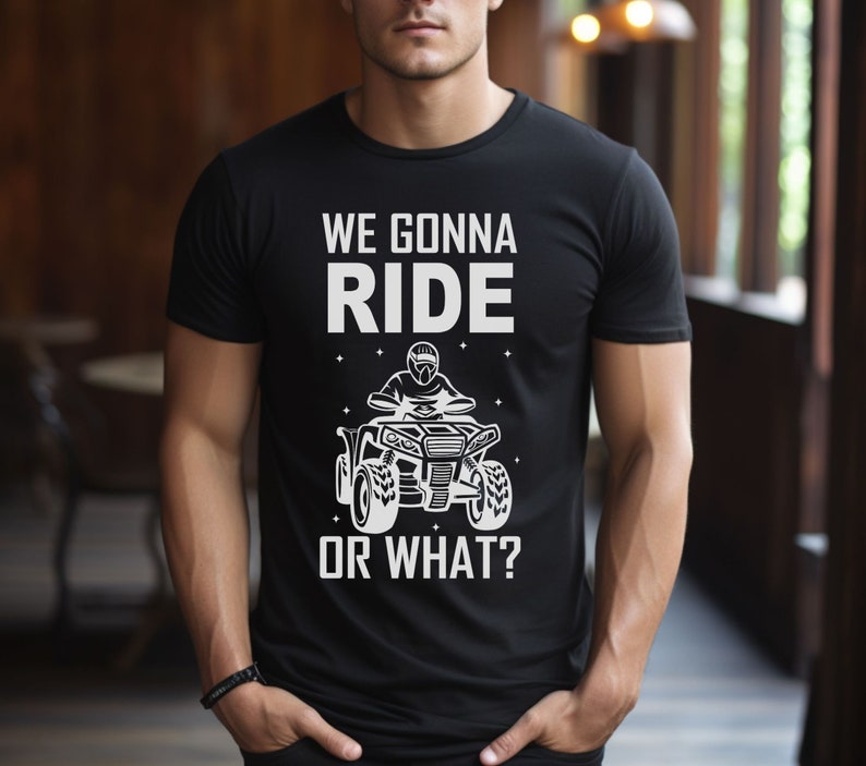 May include: Black t-shirt with a white graphic of a person riding an ATV. The text on the shirt reads "We gonna ride or what?"