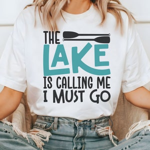 May include: White t-shirt with the text "The Lake is Calling Me I Must Go" in black and teal lettering. Two black paddles are above the text.