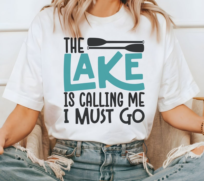 May include: White t-shirt with the text "The Lake is Calling Me I Must Go" in black and teal lettering. Two black paddles are above the text.
