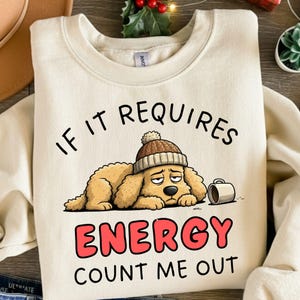 May include: Cream-colored sweatshirt with the text "IF IT REQUIRES ENERGY COUNT ME OUT". The graphic features a cartoon dog wearing a brown knit cap and looking tired, with a small coffee mug beside it.