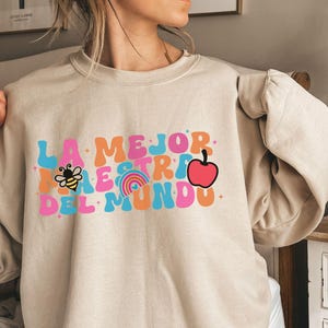 Pode incluir: Uma camisola bege com o texto "LA MEJOR MAESTRA DEL MUNDO" em letras coloridas de estilo retro. O texto inclui uma abelha, um arco-&iacute;ris e uma ma&ccedil;&atilde;.
