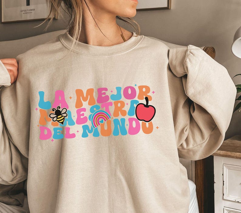 Pode incluir: Uma camisola bege com o texto "LA MEJOR MAESTRA DEL MUNDO" em letras coloridas de estilo retro. O texto inclui uma abelha, um arco-&iacute;ris e uma ma&ccedil;&atilde;.