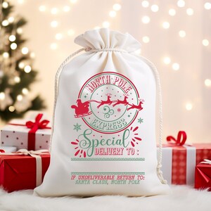 May include: White canvas gift bag with a red and green design. The bag features a Santa Claus in a sleigh pulled by reindeer with the text "North Pole Express Special Delivery To."  The bag also has the text "If undeliverable return to: Santa Claus, North Pole."