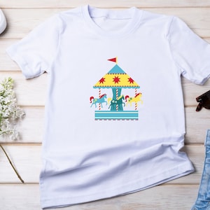 May include: White t-shirt with a colorful carousel design featuring a blue roof, yellow stars, and red and yellow horses.