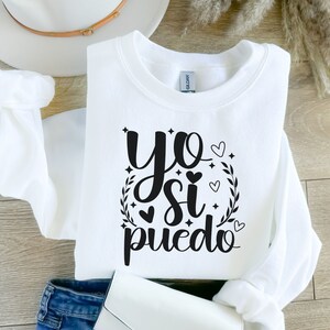 May include: White sweatshirt with black text that reads "yo si puedo" with a wreath and hearts around the text.