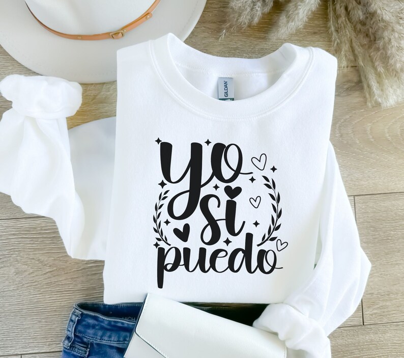 May include: White sweatshirt with black text that reads "yo si puedo" with a wreath and hearts around the text.