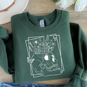 May include: A dark green sweatshirt with a white graphic of a skull with flowers growing out of the top. The skull is outlined in a white square.