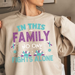 以下が含まれることがあります： ベージュのトレーナーに、「In this family no one fights alone」という文字が紫とターコイズブルーで書かれています。文字の周りは、スタイリッシュなターコイズブルーの花と紫のリボンで囲まれています。