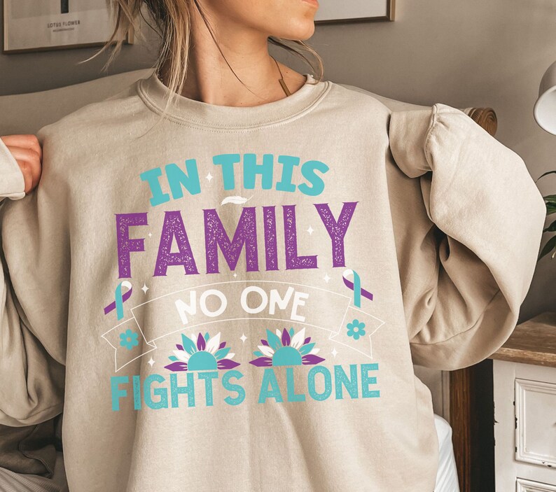 以下が含まれることがあります： ベージュのトレーナーに、「In this family no one fights alone」という文字が紫とターコイズブルーで書かれています。文字の周りは、スタイリッシュなターコイズブルーの花と紫のリボンで囲まれています。