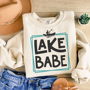 May include: A cream-colored sweatshirt with a blue and white graphic that says "LAKE BABE" with a silhouette of a person in a canoe.