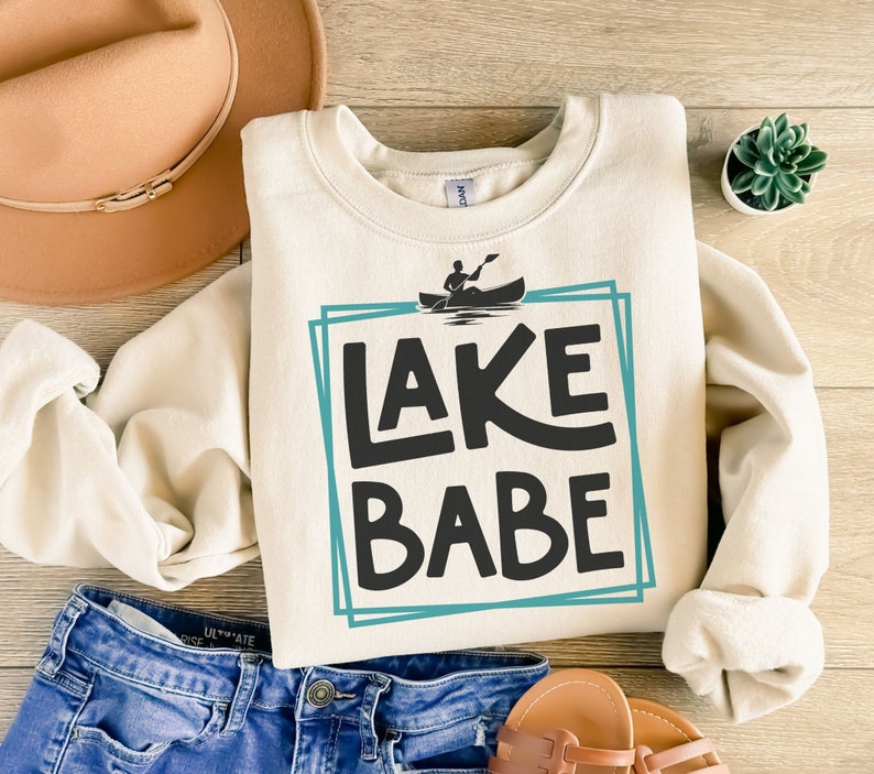 May include: A cream-colored sweatshirt with a blue and white graphic that says "LAKE BABE" with a silhouette of a person in a canoe.