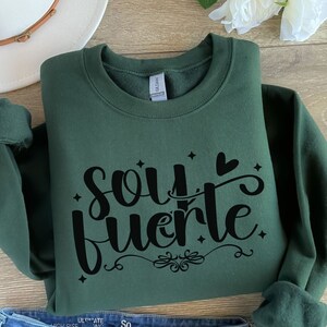 May include: A dark green sweatshirt with the text "soy fuerte" in black lettering. The text is in a stylized font with a heart and stars.
