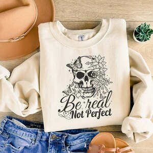 May include: A cream-colored sweatshirt with a black and white graphic of a skull with flowers and butterflies. The text "Be real Not Perfect" is printed below the skull.
