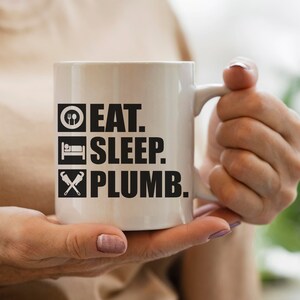 May include: White ceramic mug with black text that reads "Eat. Sleep. Plumb."  The mug has a black and white icon of a fork and spoon, a bed, and a wrench and pipe.