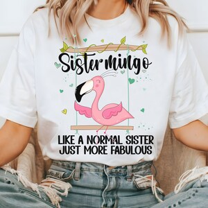 Puede incluir: Camiseta blanca con un flamenco rosa en un columpio y el texto "Sistermingo Like a normal sister just more fabulous".