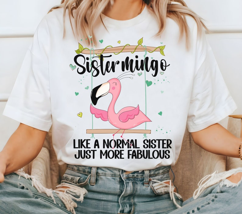 Puede incluir: Camiseta blanca con un flamenco rosa en un columpio y el texto "Sistermingo Like a normal sister just more fabulous".