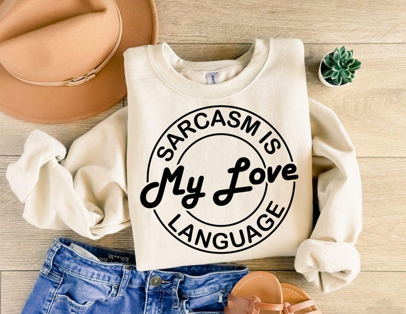 K&ouml;nnte beinhalten: Ein cremefarbener Pullover mit einem schwarzen Kreis-Motiv, auf dem "Sarkasmus ist meine Liebessprache" steht.