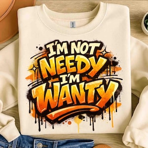 Pu&ograve; includere: Felpa color crema con la scritta "I'M NOT NEEDY I'M WANTY" in un design in stile graffiti. Il testo &egrave; arancione e giallo con contorni e gocce nere. La felpa &egrave; piegata su una superficie di legno.