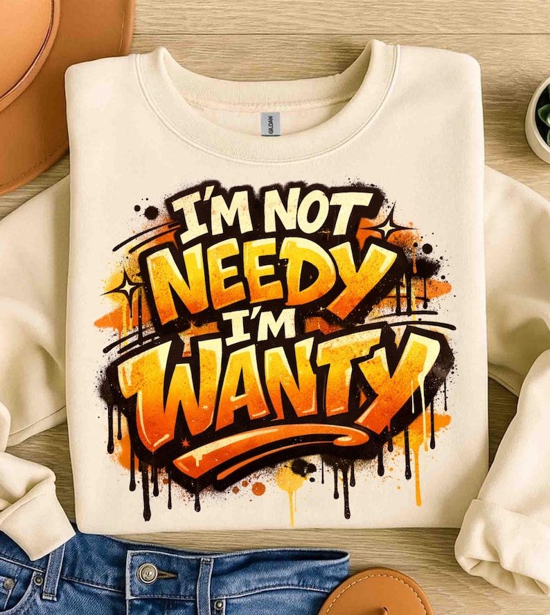 Pu&ograve; includere: Felpa color crema con la scritta "I'M NOT NEEDY I'M WANTY" in un design in stile graffiti. Il testo &egrave; arancione e giallo con contorni e gocce nere. La felpa &egrave; piegata su una superficie di legno.