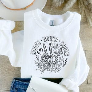 May include: White sweatshirt with a black graphic design of a skeleton hand making a peace sign with the words "BOOKS" on either side of the hand. The graphic also includes butterflies, flowers, and lightning bolts.