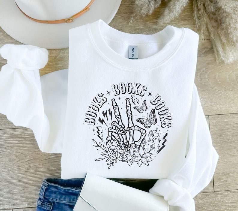 May include: White sweatshirt with a black graphic design of a skeleton hand making a peace sign with the words "BOOKS" on either side of the hand. The graphic also includes butterflies, flowers, and lightning bolts.