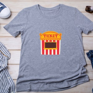 May include: Gray t-shirt with a graphic of a red and white striped ticket booth with the word "TICKET" in yellow letters.