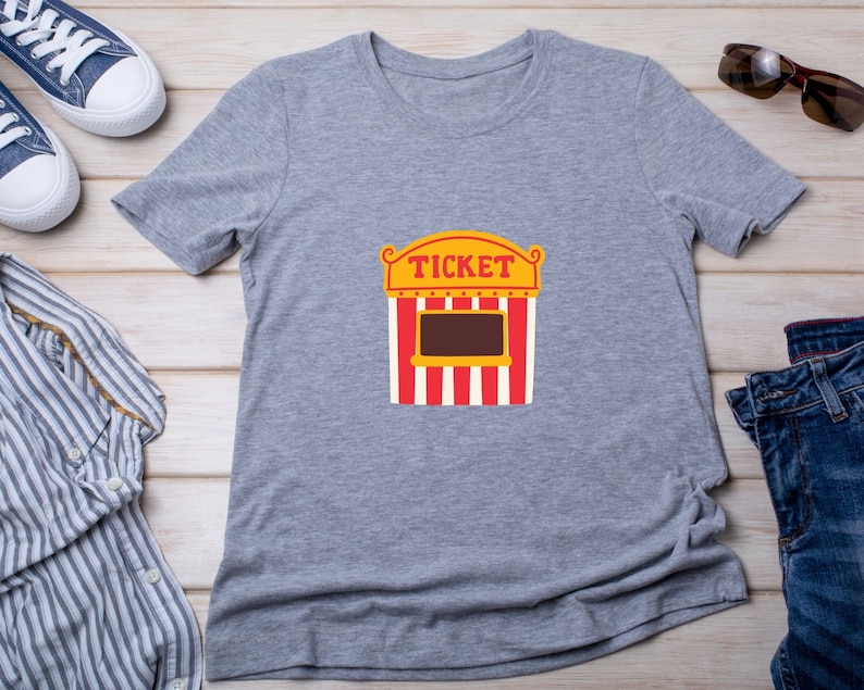 May include: Gray t-shirt with a graphic of a red and white striped ticket booth with the word "TICKET" in yellow letters.