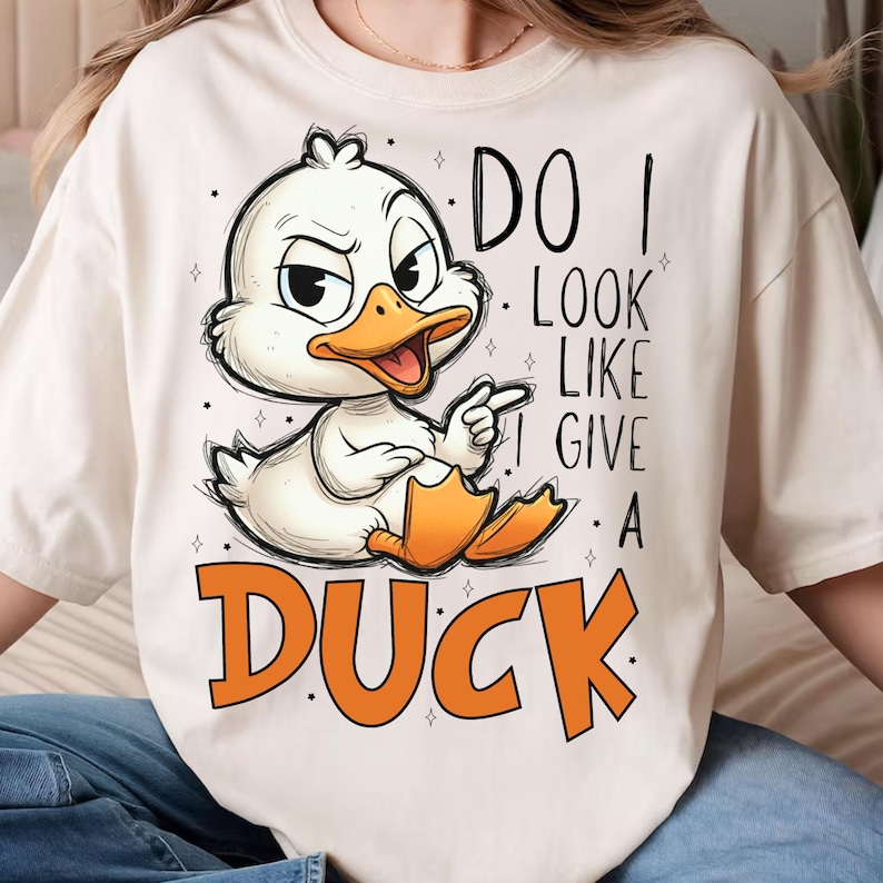 May include: Cream-colored t-shirt featuring a cartoon duck with an annoyed expression. The duck is white with an orange beak and feet, pointing with one wing. Text reads "DO I LOOK LIKE I GIVE A DUCK" in black and orange.